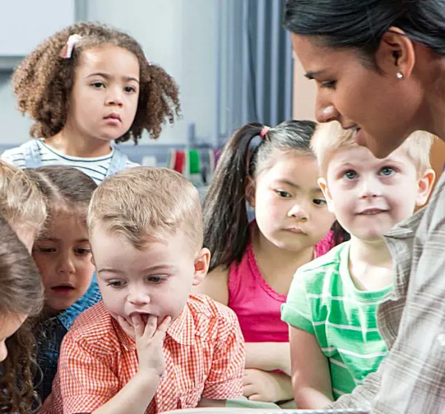 A lady reads story from a storybook while the children gather around to listen to the story